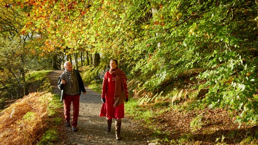 Two visitors in warm coats walk along a lakeshore path by Derwent Water, with autumnal leaves turning on the trees